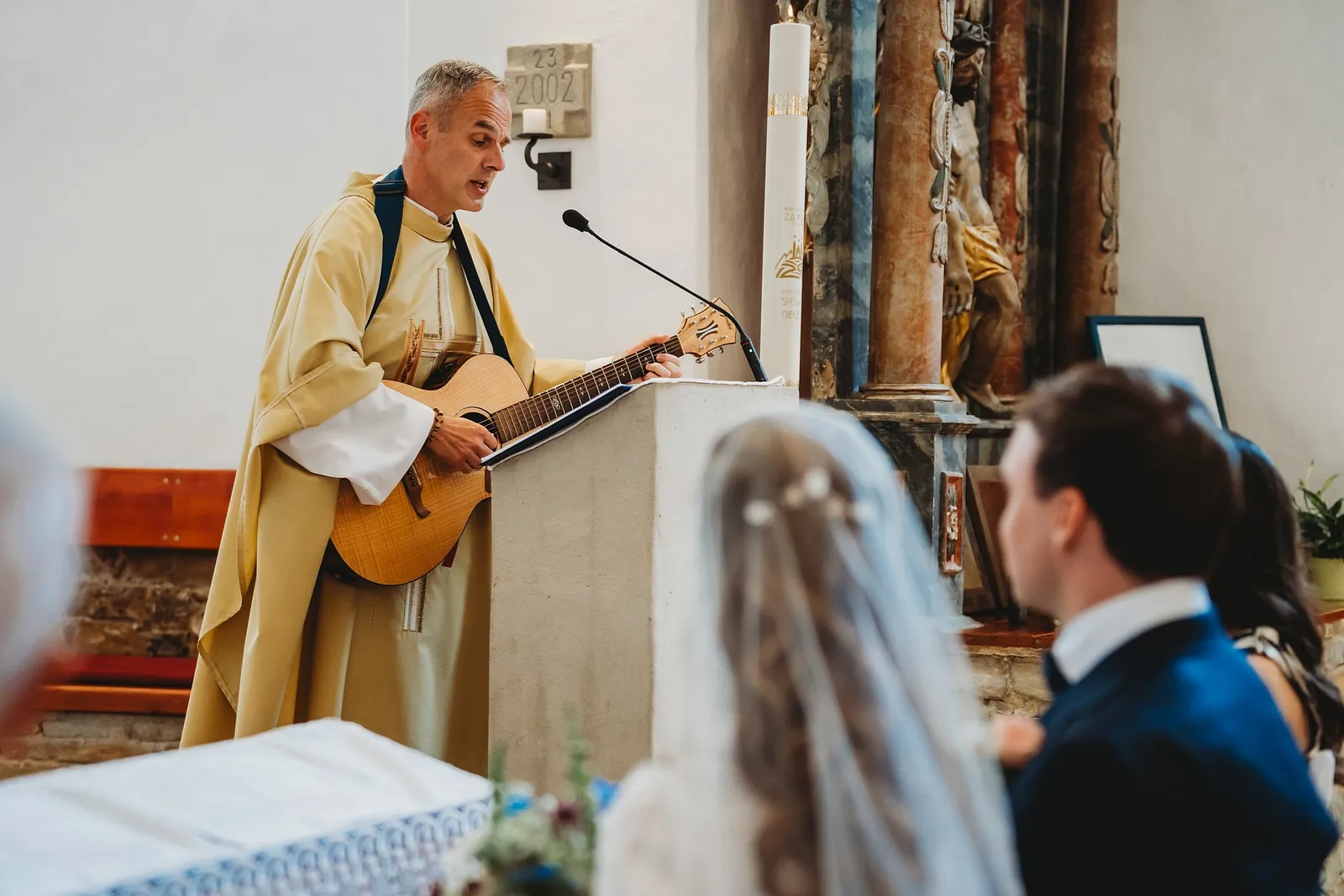 Kňaz hrá na gitare pri ambóne, nevesta v závoji a ženích ho sediac počúvajú – svadba Sandra a Sebastian, Stodola Gréta