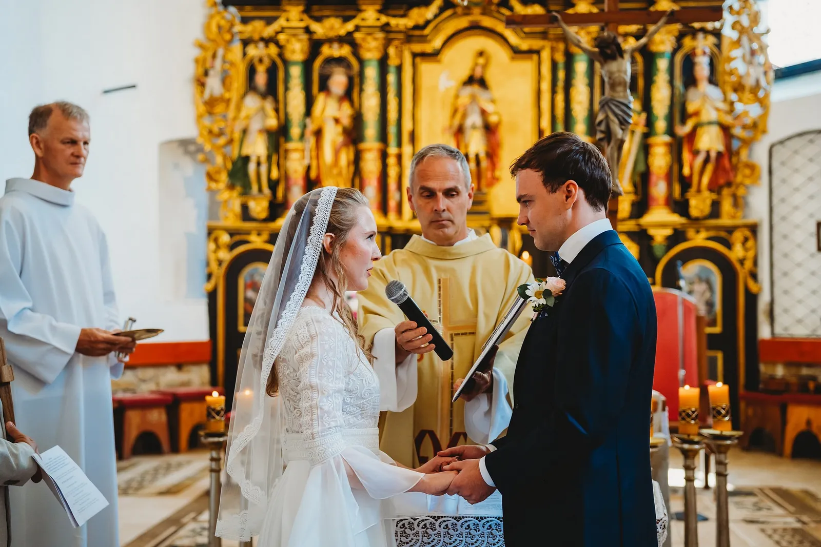 Nevesta vyslovuje sľub do mikrofónu a drží ženícha za ruky pred kňazom a zlatým oltárom – svadba Sandra a Sebastian, Stodola Gréta