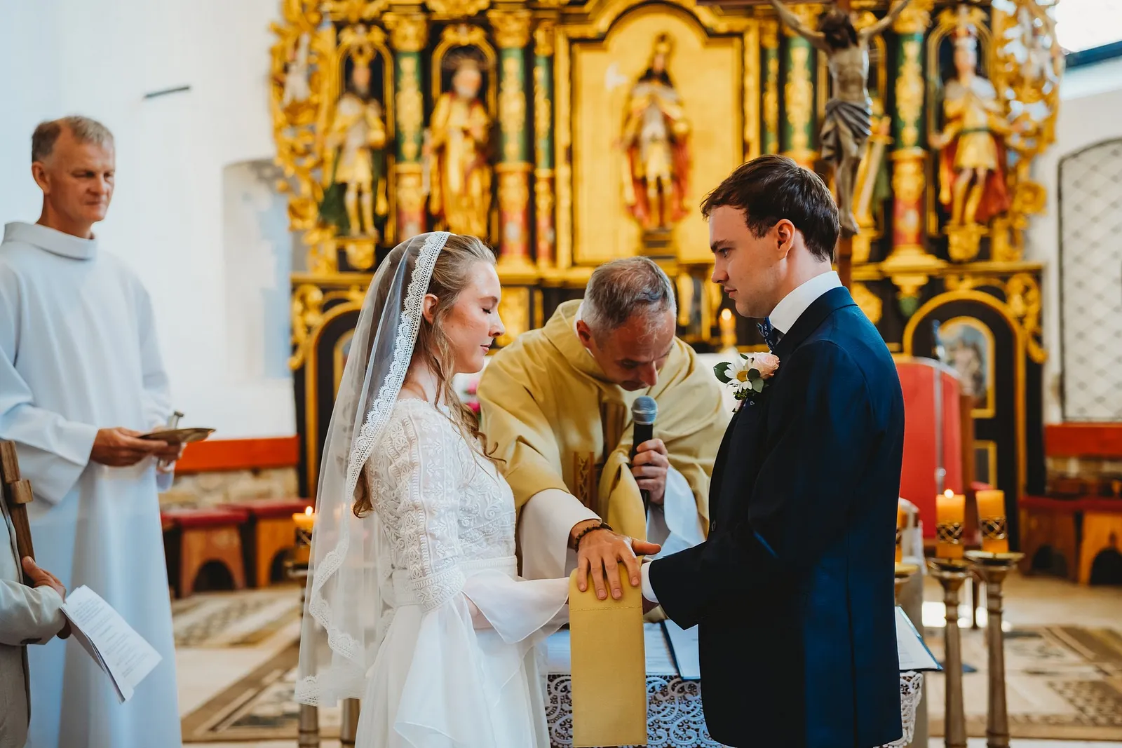 Kňaz zväzuje ruky páru štólou pri oltári, vedľa stojí muž v albe s miskou na obrúčky – svadba Sandra a Sebastian, Stodola Gréta