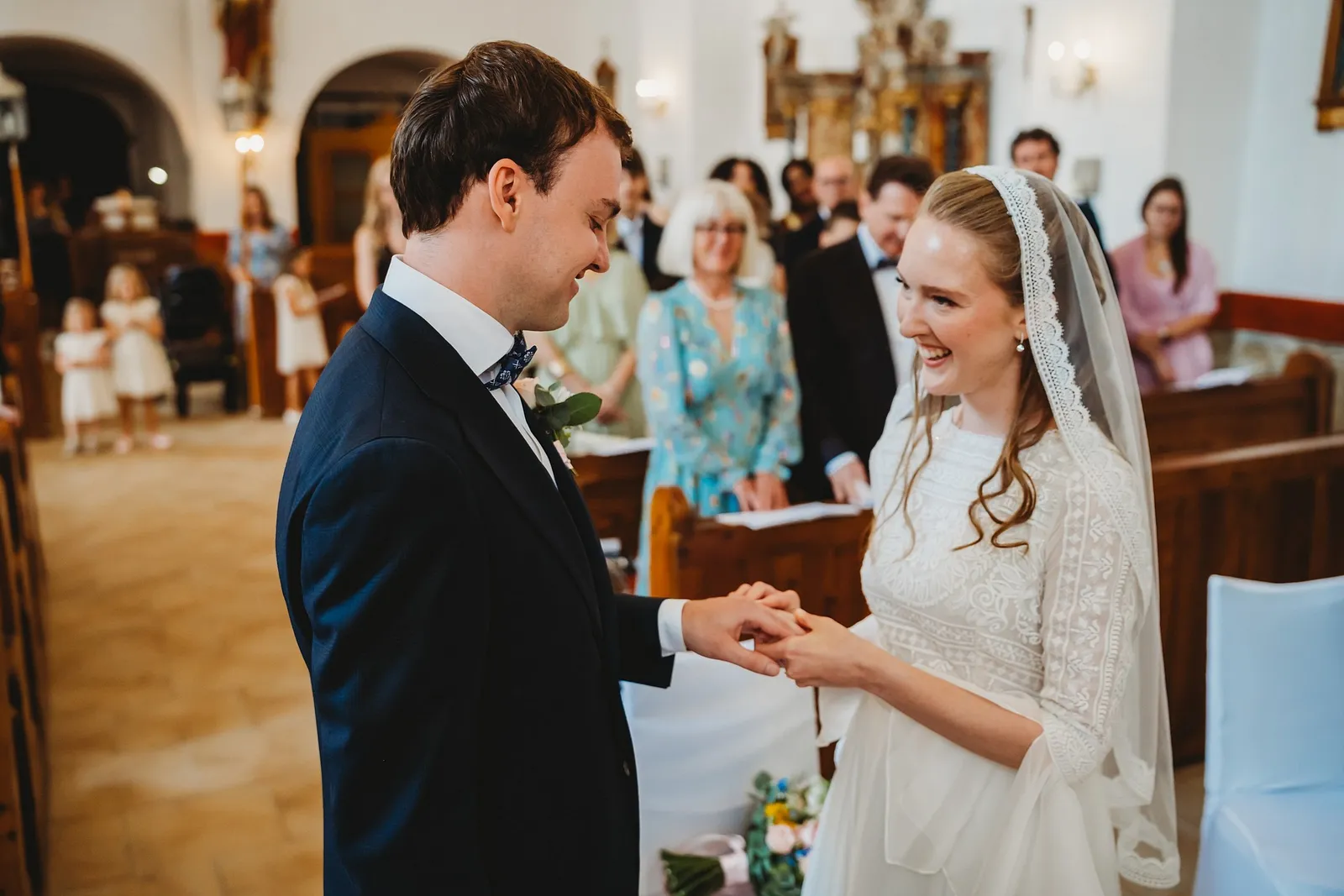 Rozžiarená nevesta navlieka ženíchovi obrúčku, v uličke kostola stoja malé dievčatá v bielom – svadba Sandra a Sebastian, Stodola Gréta