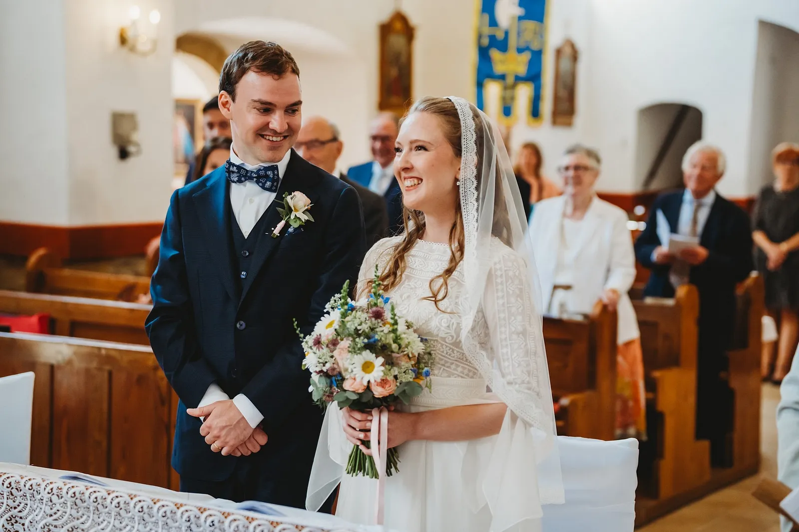 Usmiaty ženích s motýlikom a nevesta s lúčnou kyticou stoja pred oltárom v kostole – svadba Sandra a Sebastian, Stodola Gréta
