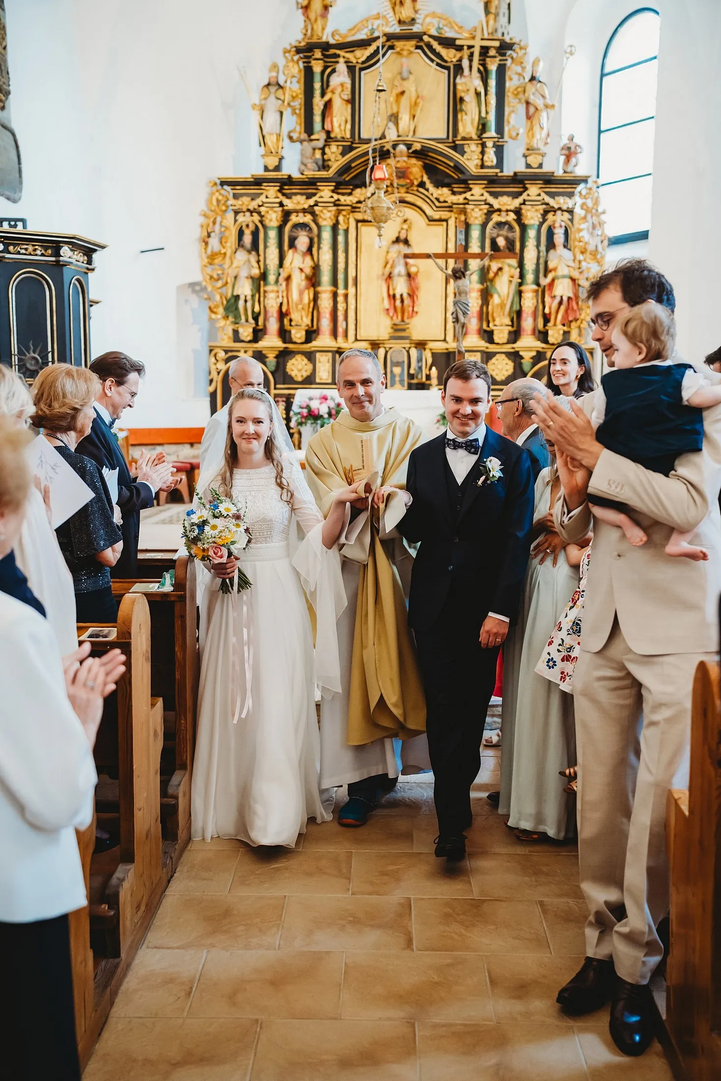 Novomanželia s kňazom odchádzajú uličkou kostola pred zlatým oltárom, hostia tlieskajú – svadba Sandra a Sebastian, Stodola Gréta
