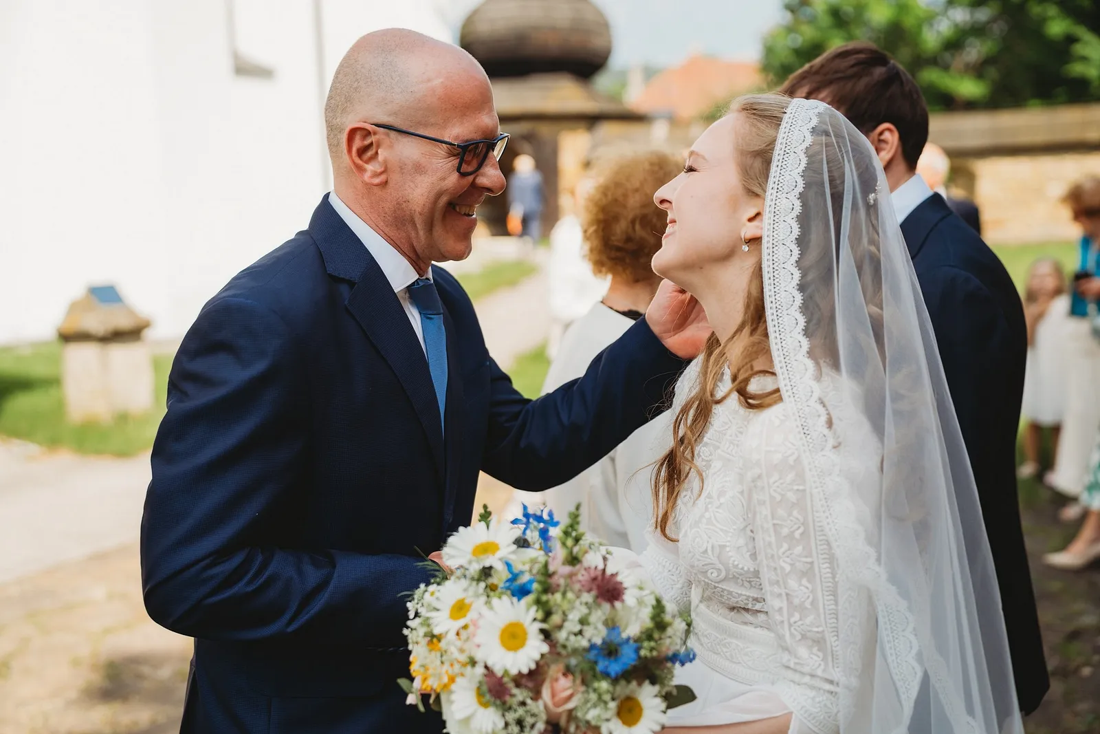 Hosť v okuliaroch láskavo hladí nevestu po tvári pri gratulácii pred kostolom – svadba Sandra a Sebastian, Stodola Gréta