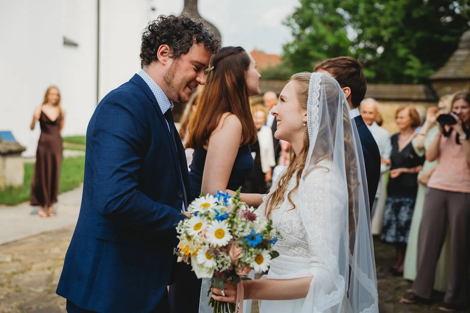 Kučeravý hosť s úsmevom gratuluje neveste v závoji, v pozadí čakajú ďalší hostia – svadba Sandra a Sebastian, Stodola Gréta