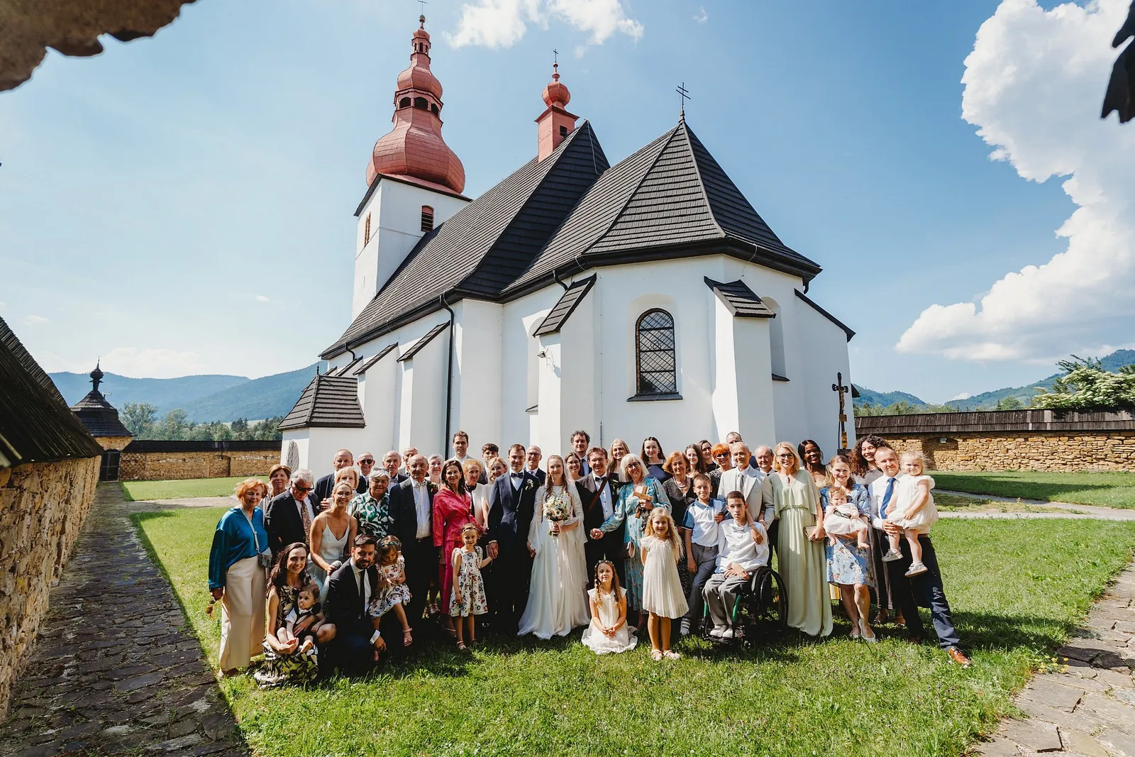 Spoločná fotka svadobných hostí na tráve pred bielym kostolom s červenými vežami a horami – svadba Sandra a Sebastian, Stodola Gréta