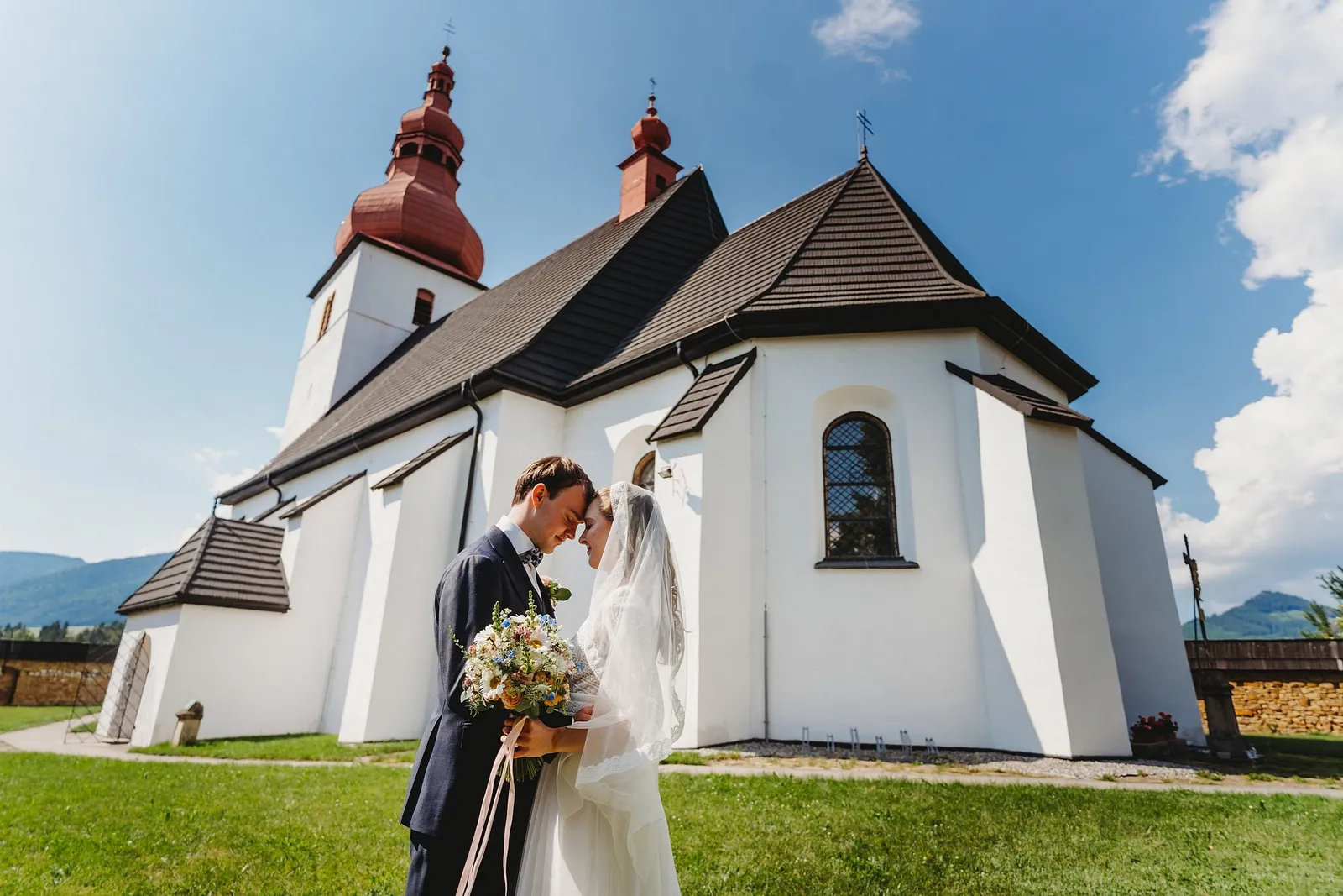 Novomanželia sa objímajú pred bielym kostolom s červenými vežami pod modrou oblohou – svadba Sandra a Sebastian, Stodola Gréta