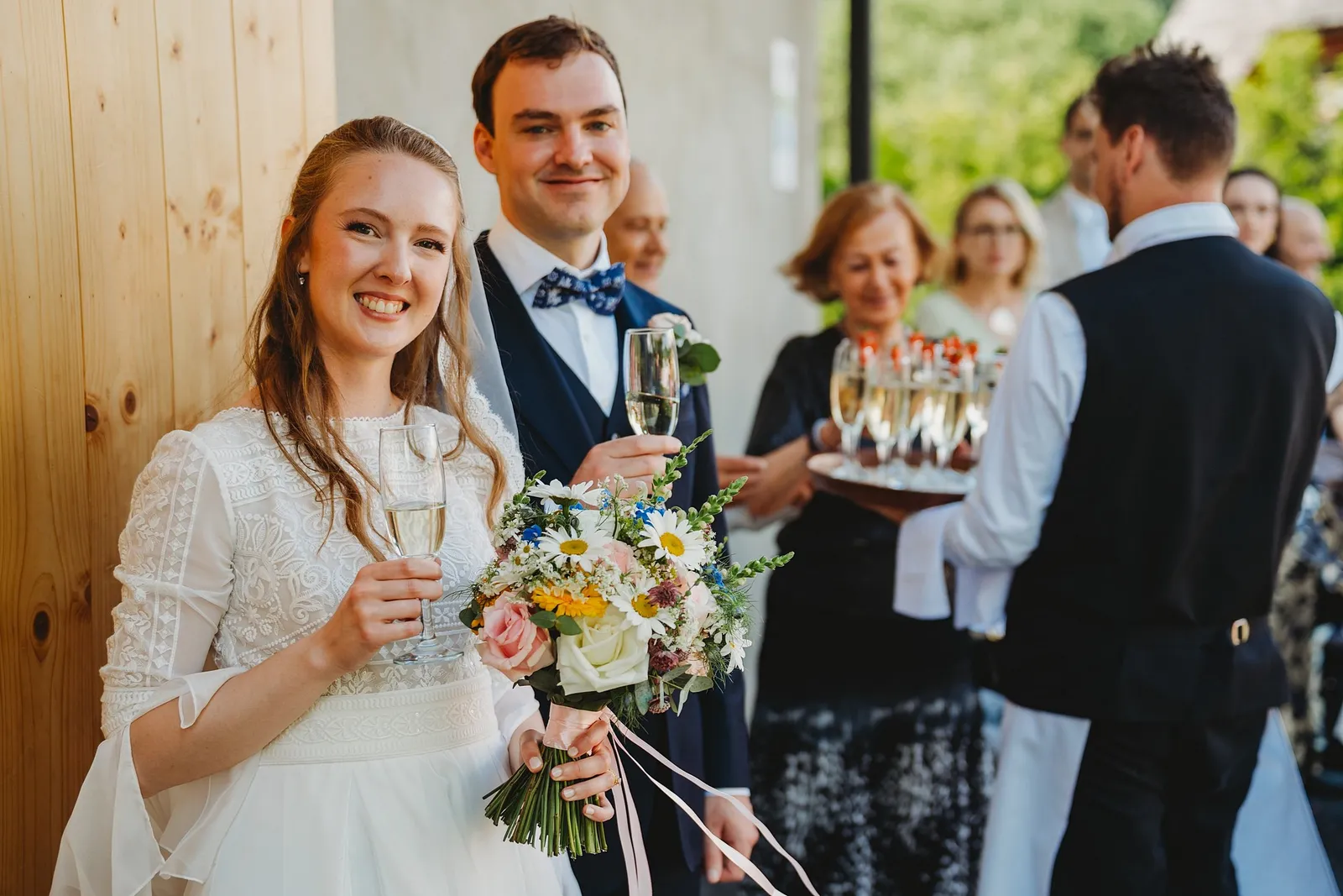 Usmiata nevesta s kyticou a ženích s pohárom šampanského pri privítaní, čašník nesie podnos – svadba Sandra a Sebastian, Stodola Gréta