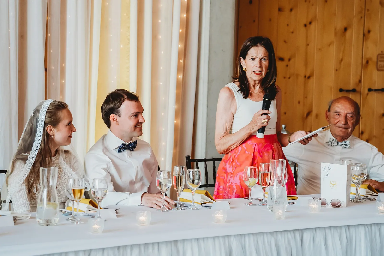 Žena v ružovej sukni má príhovor s mikrofónom pri hlavnom stole s novomanželmi – svadba Sandra a Sebastian, Stodola Gréta
