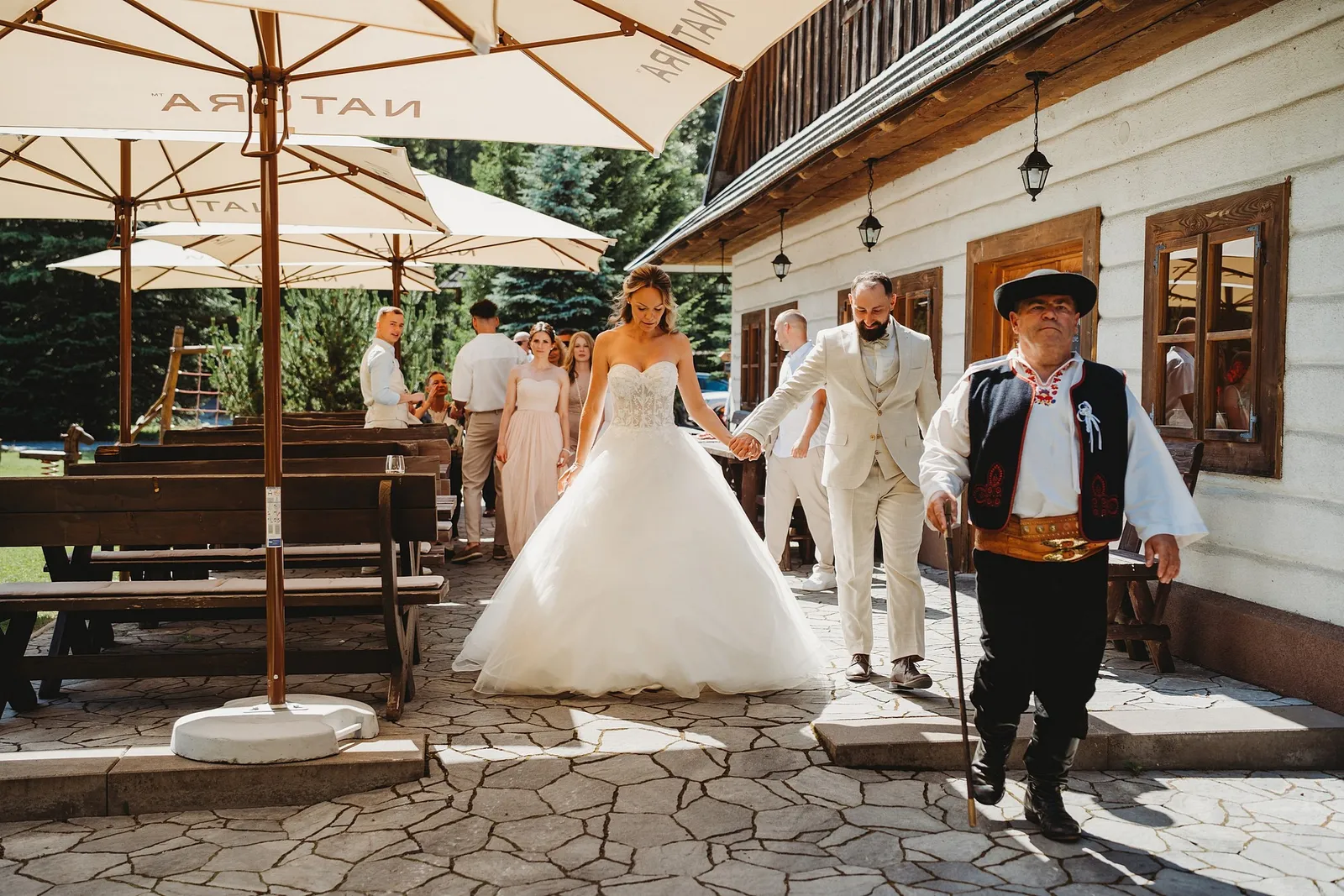 Starejší v kroji s valaškou vedie novomanželov cez terasu so slnečníkmi – svadba Simonka a Nuri, Stodola Liptovský dvor