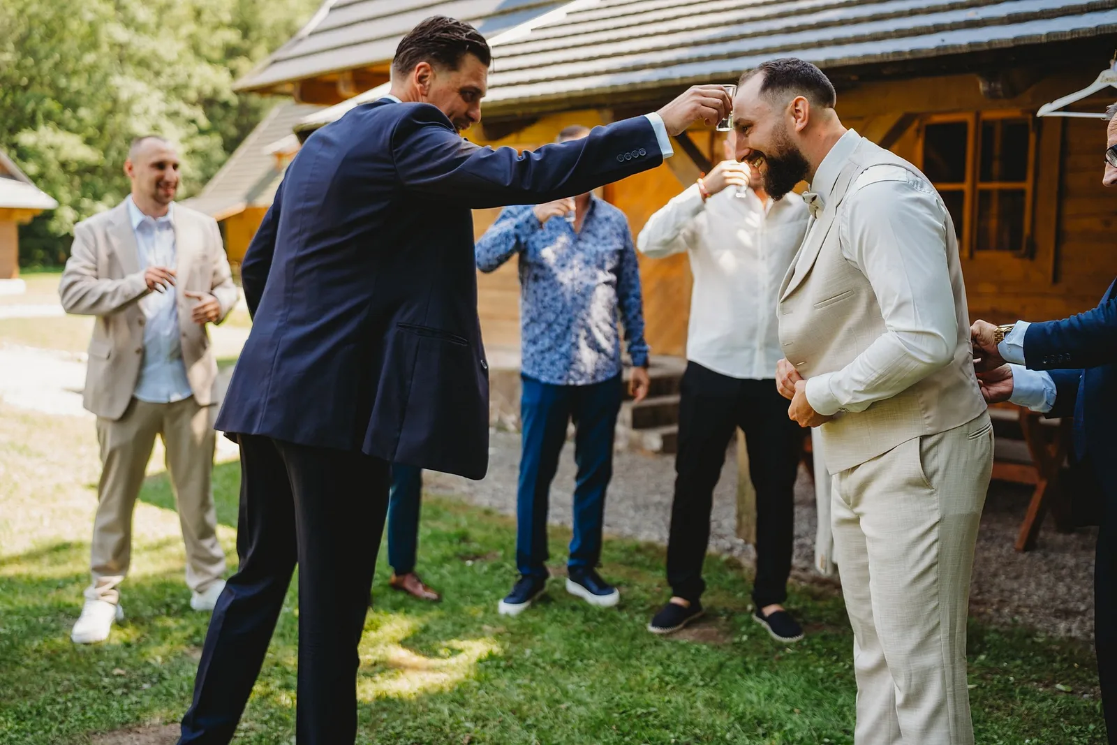 Ženích v béžovom obleku vypíja štamperlík, kamarát mu ho drží nad hlavou – svadba Simonka a Nuri, Stodola Liptovský dvor