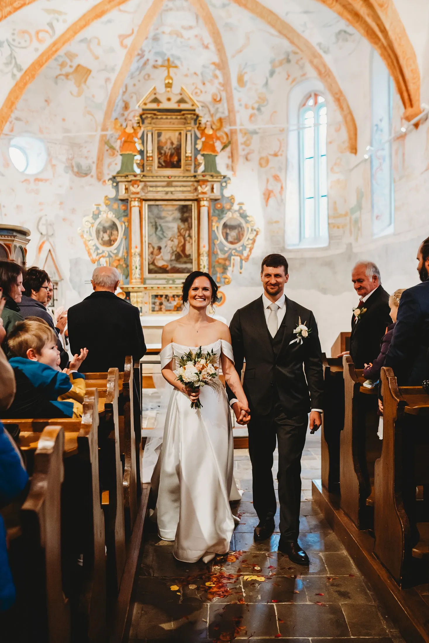 Novomanželia odchádzajú uličkou kostola s maľovanou klenbou a oltárom, na zemi lupene – svadba Andrejka a Janko, Stodola Liptovský dvor