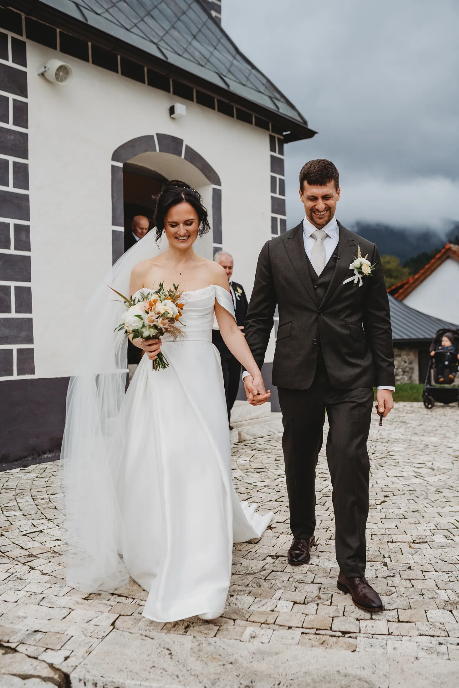 Novomanželia vychádzajú z bieleho kostolíka na dláždené nádvorie, za nimi hory v oblakoch – svadba Andrejka a Janko, Stodola Liptovský dvor
