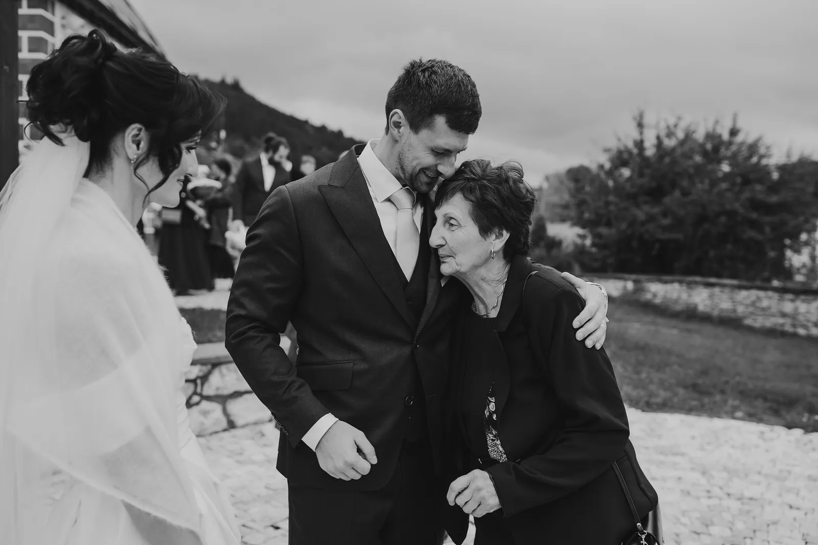 Čiernobiele objatie ženícha so staršou ženou pred kostolom, vedľa stojí nevesta so závojom – svadba Andrejka a Janko, Stodola Liptovský dvor