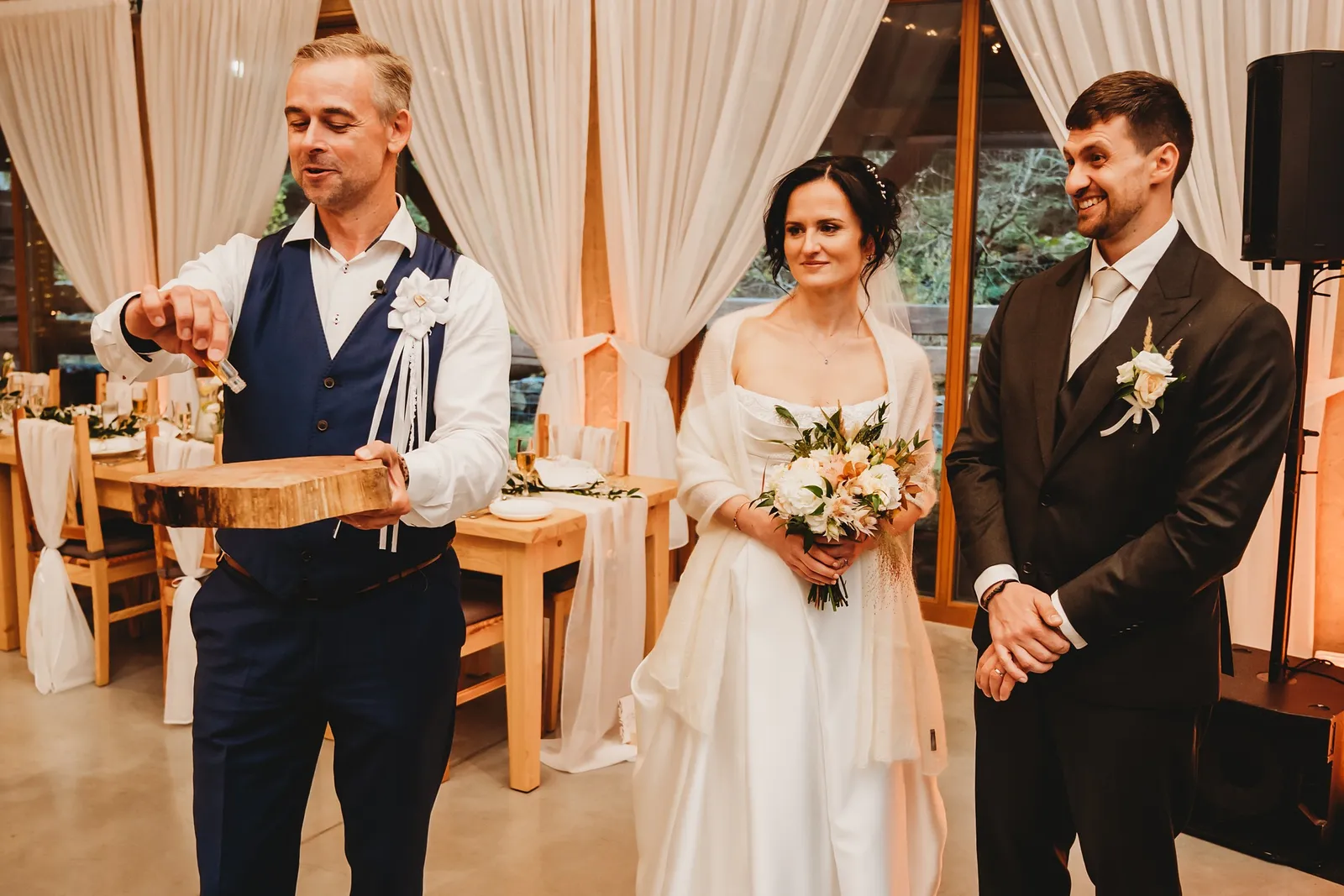 Muž vo veste s pierkom drží drevený podnos, vedľa stojí nevesta s kyticou a ženích – svadba Andrejka a Janko, Stodola Liptovský dvor