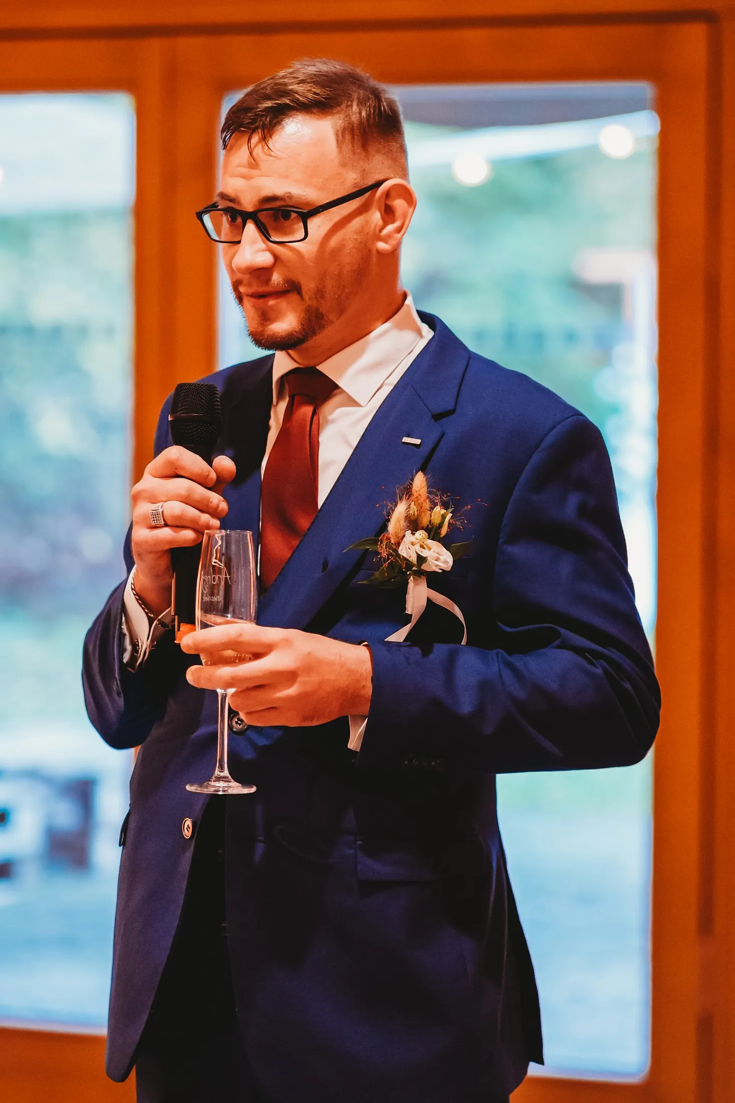 Muž v modrom obleku s okuliarmi hovorí príhovor s mikrofónom a pohárom šampanského – svadba Andrejka a Janko, Stodola Liptovský dvor