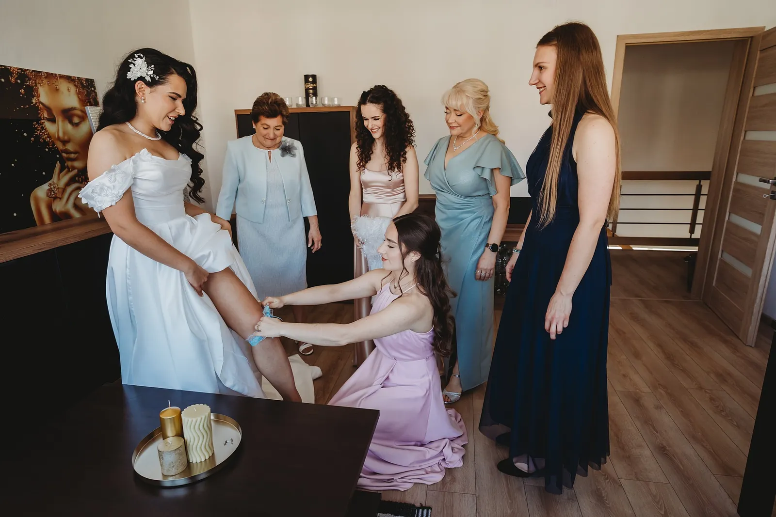 Družička v ružových šatách navlieka neveste podväzok, mama a ďalšie ženy sa usmievajú – svadba Denisa a Martin, Stodola na poli