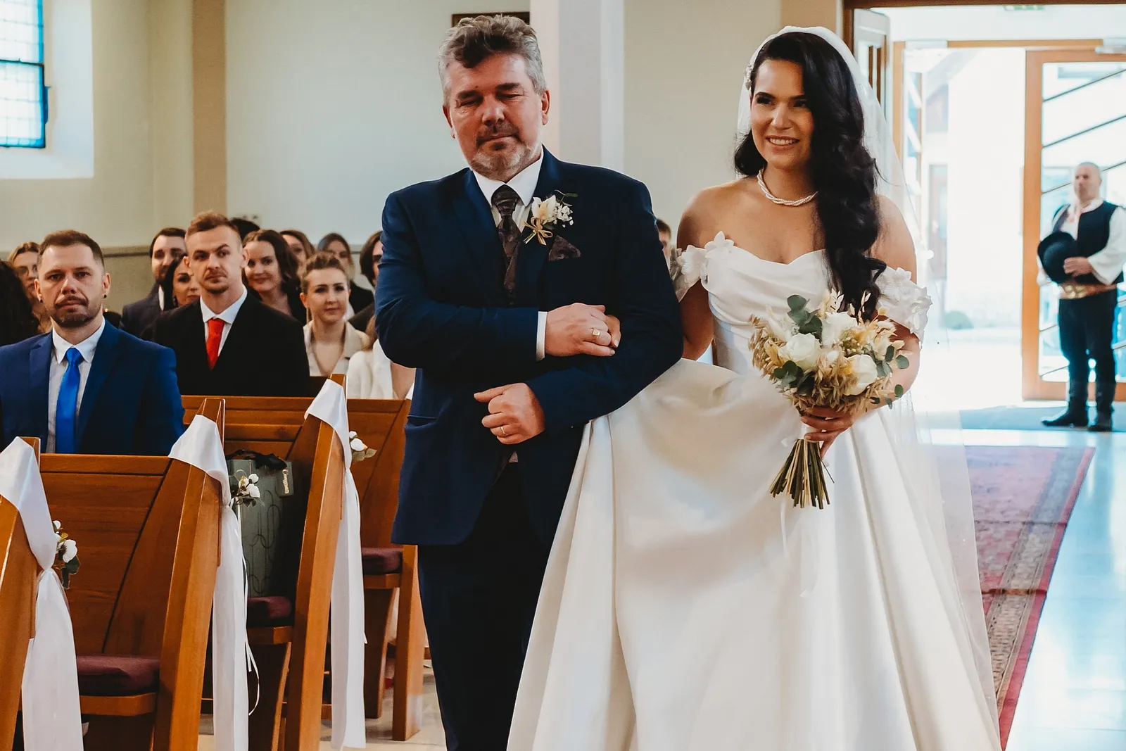 Otec vedie usmiatu nevestu s kyticou uličkou kostola k oltáru, hostia sa otáčajú – svadba Denisa a Martin, Stodola na poli