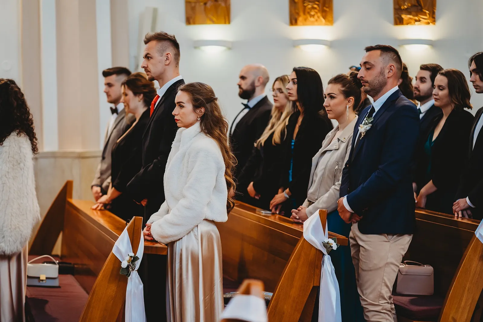 Svadobní hostia v elegantnom oblečení stoja v laviciach kostola počas obradu – svadba Denisa a Martin, Stodola na poli