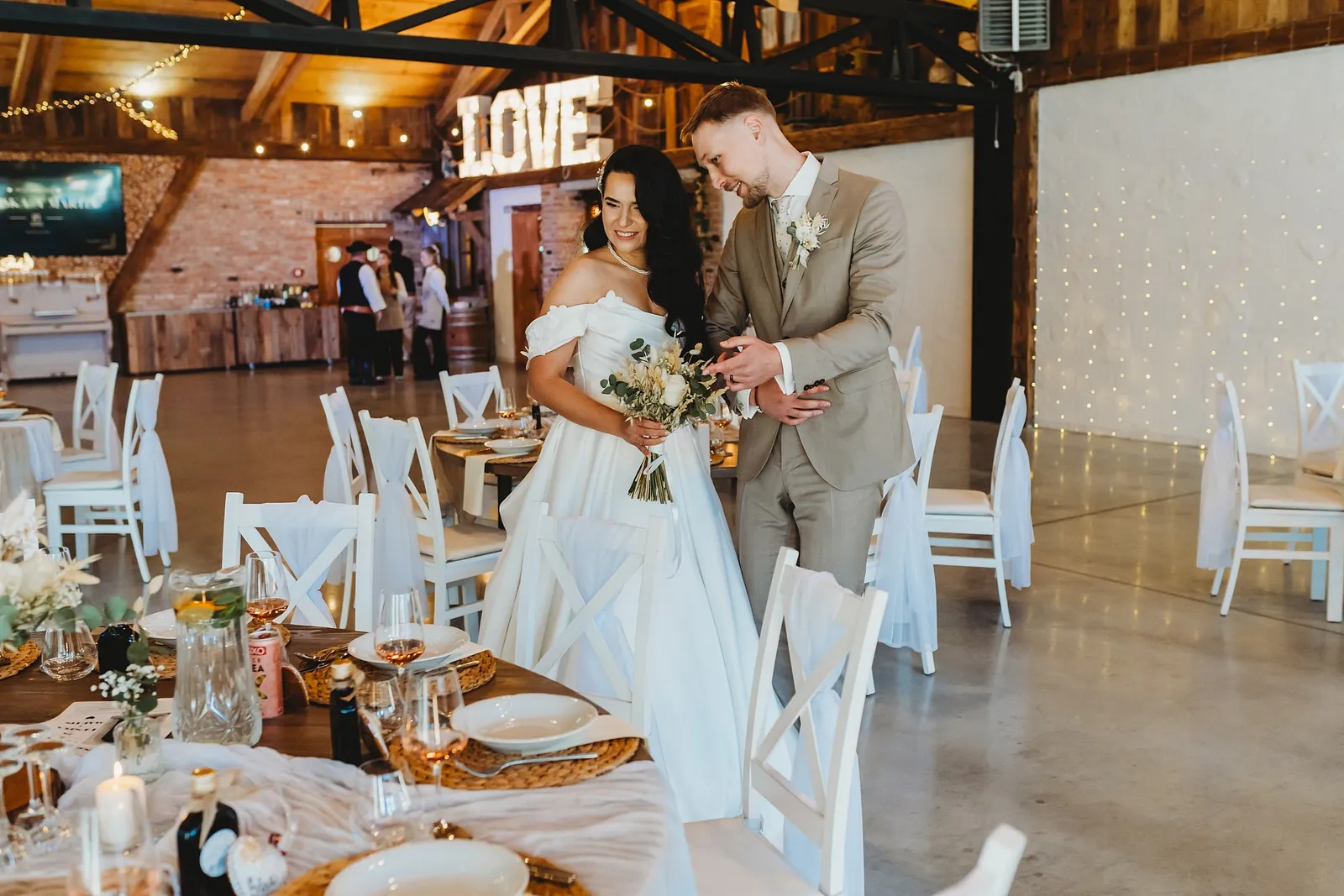 Nevesta s kyticou a ženích si prezerajú prestreté stoly pod svetelným nápisom LOVE – svadba Denisa a Martin, Stodola na poli