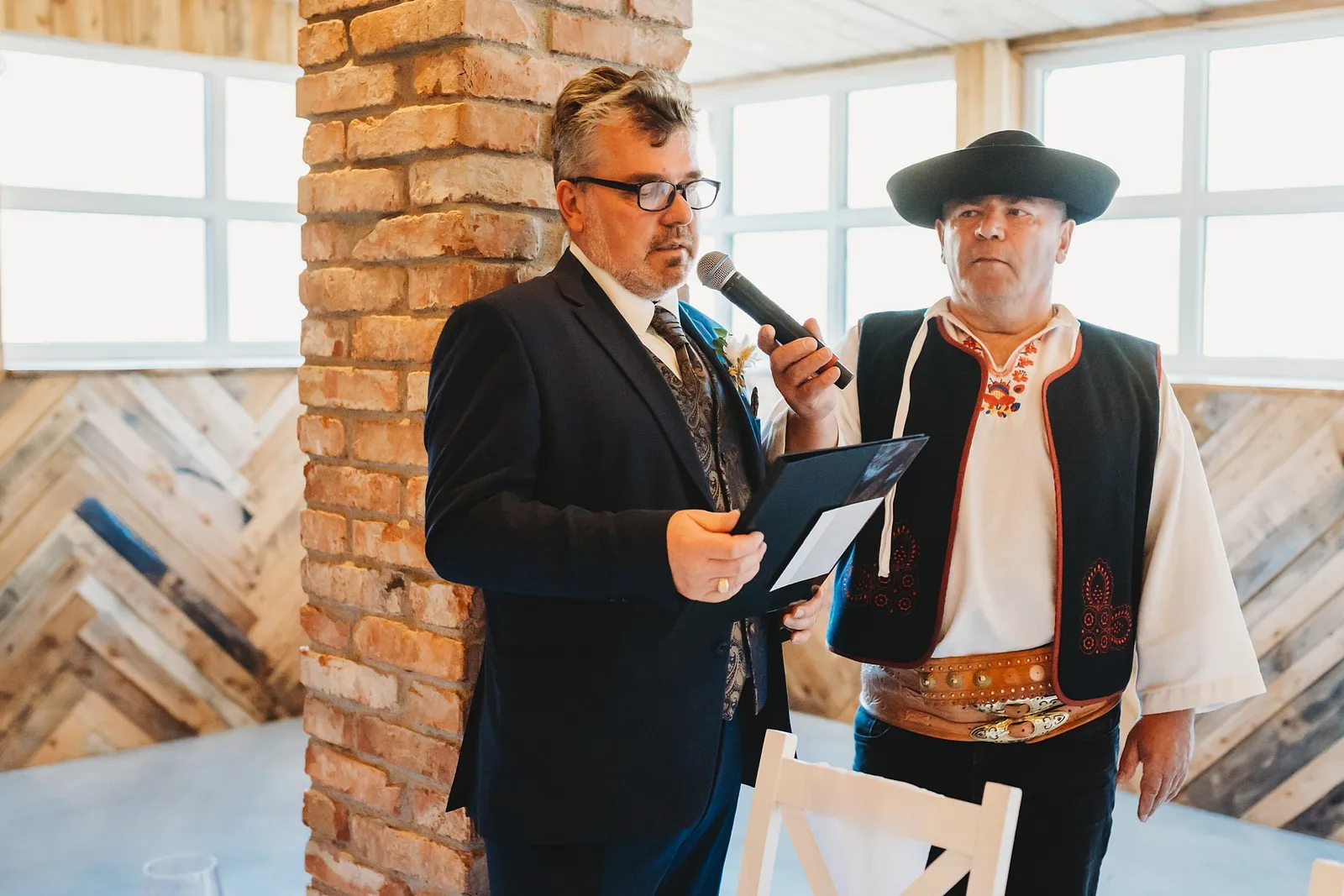 Muž v tmavom obleku hovorí do mikrofónu príhovor, vedľa neho stojí muž v ľudovom kroji a klobúku – svadba Denisa a Martin, Stodola na poli