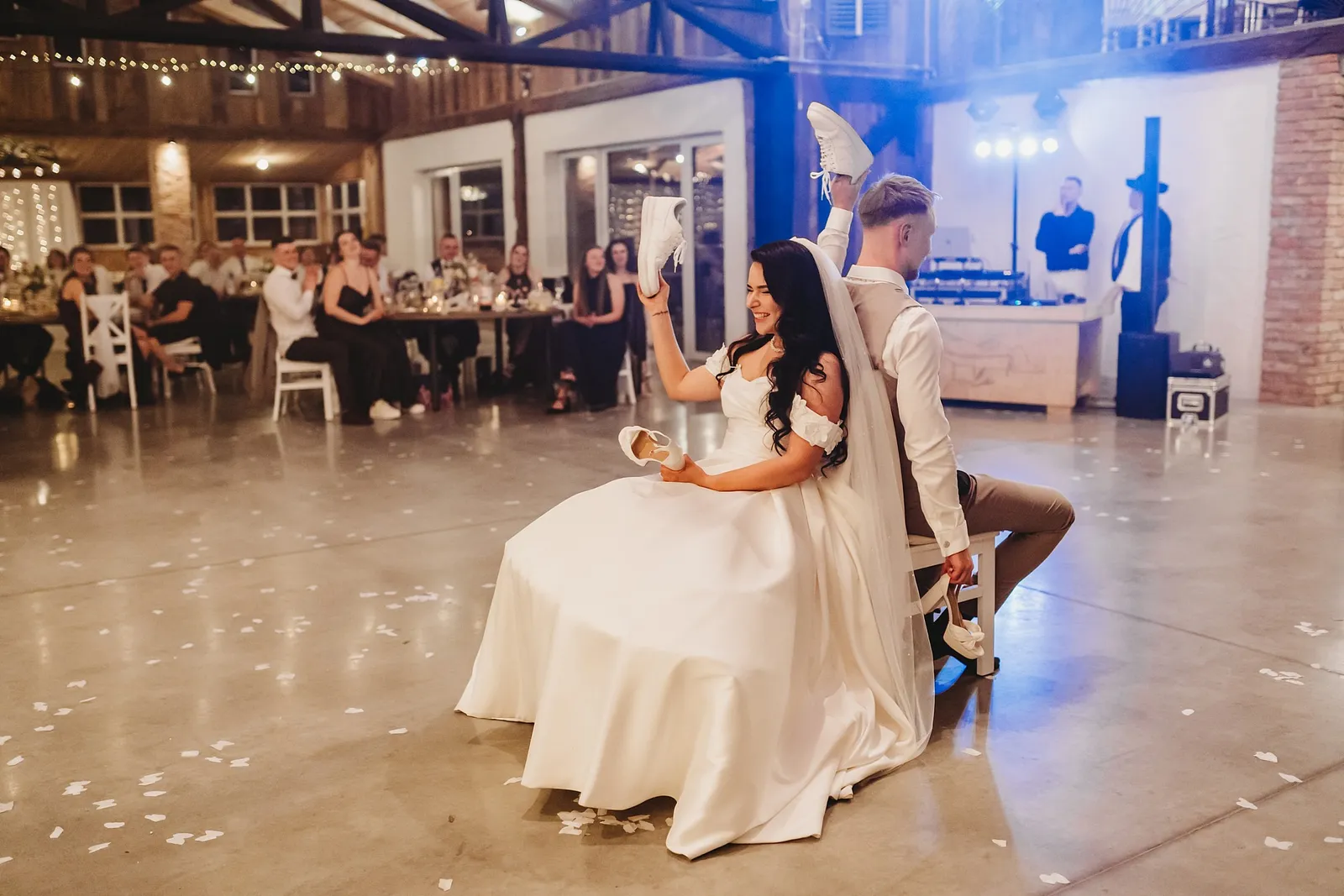 Nevesta a ženích sedia chrbtom k sebe a zdvíhajú topánky pri svadobnej hre o manželstve – svadba Denisa a Martin, Stodola na poli