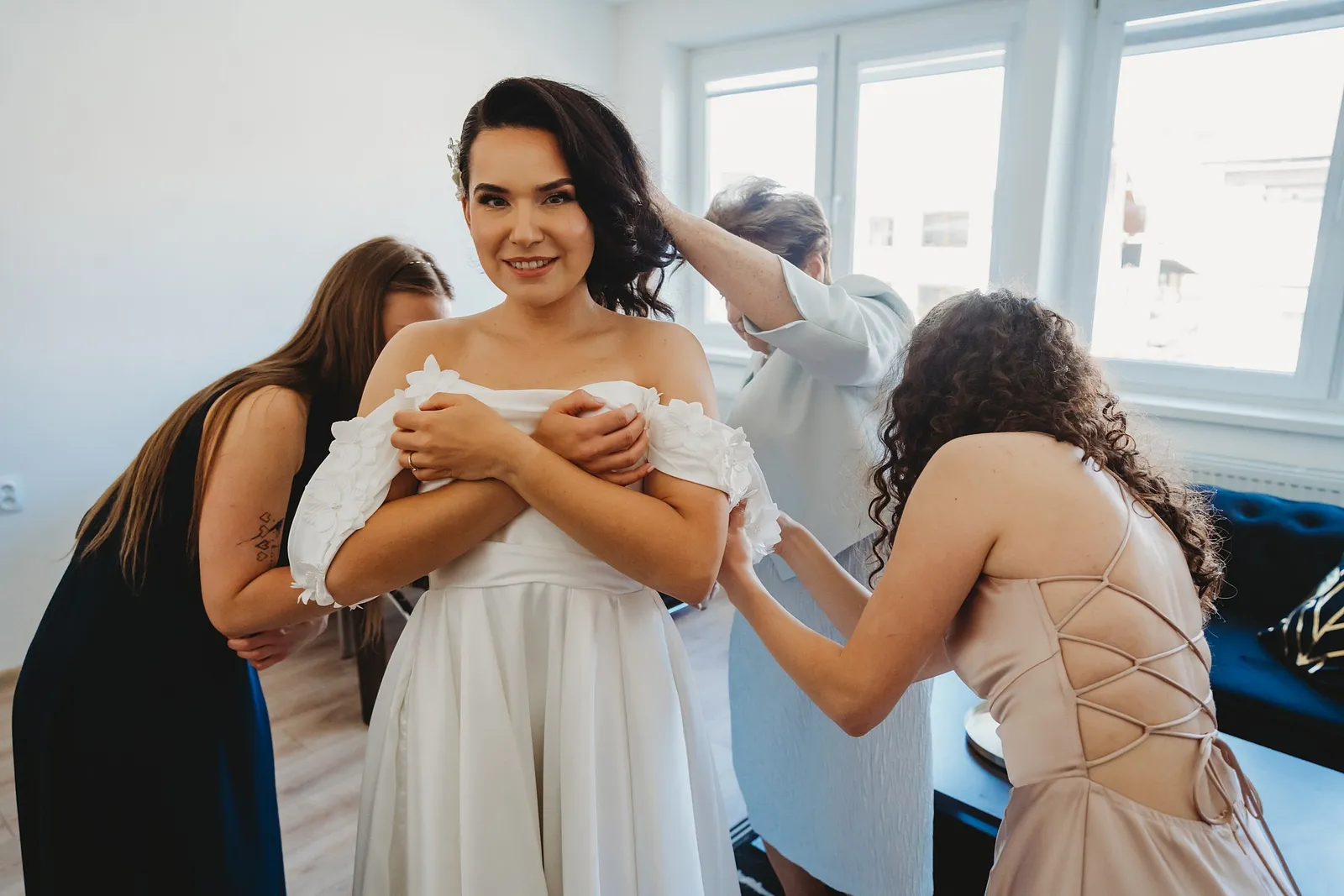Družičky a mama pomáhajú neveste zapnúť biele šaty s odhalenými ramenami pri okne – svadba Denisa a Martin, Stodola na poli
