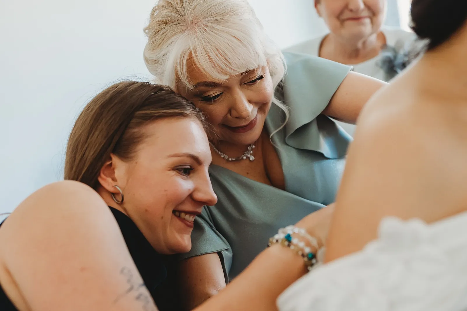 Dojatá mama a družička sa s úsmevom túlia pri neveste počas obliekania svadobných šiat – svadba Denisa a Martin, Stodola na poli