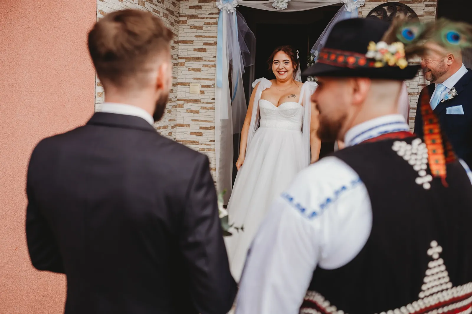 Usmiata nevesta vychádza z dverí ozdobených tylom, víta ju ženích a starejší v kroji – svadba Natália a Tomáš, Hotel Demänová