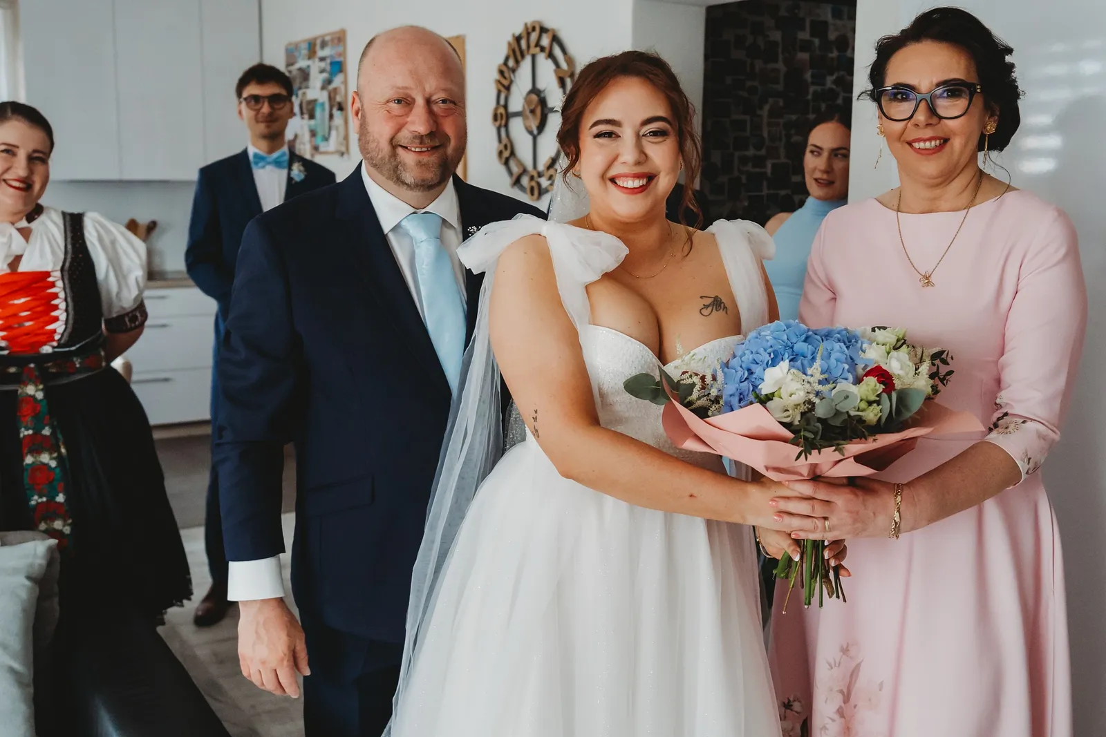 Žiariaca nevesta s kyticou modrých hortenzií pózuje medzi rodičmi, za nimi hostia – svadba Natália a Tomáš, Hotel Demänová