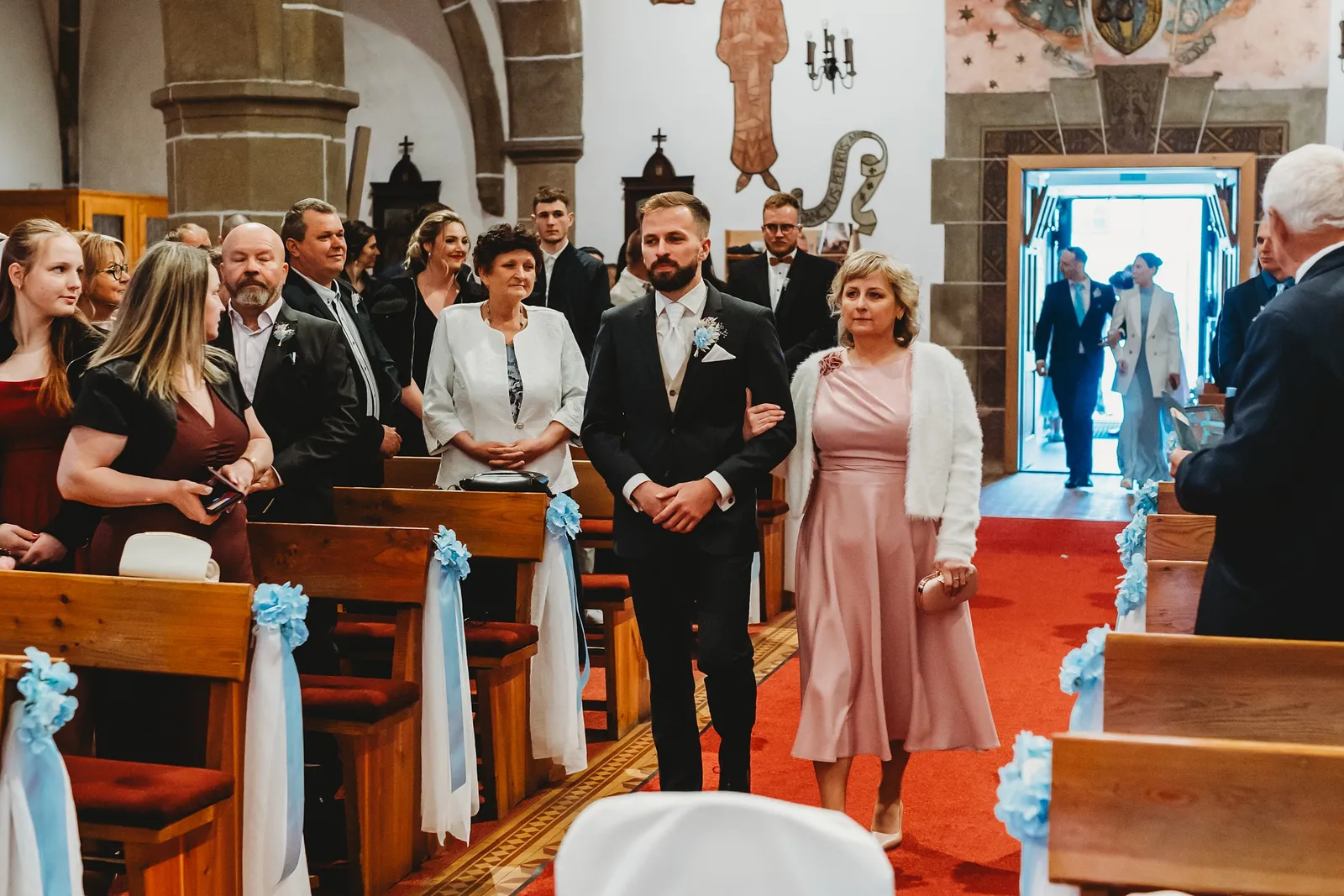Ženích kráča s mamou uličkou kostola okolo lavíc ozdobených modrými mašľami – svadba Natália a Tomáš, Hotel Demänová