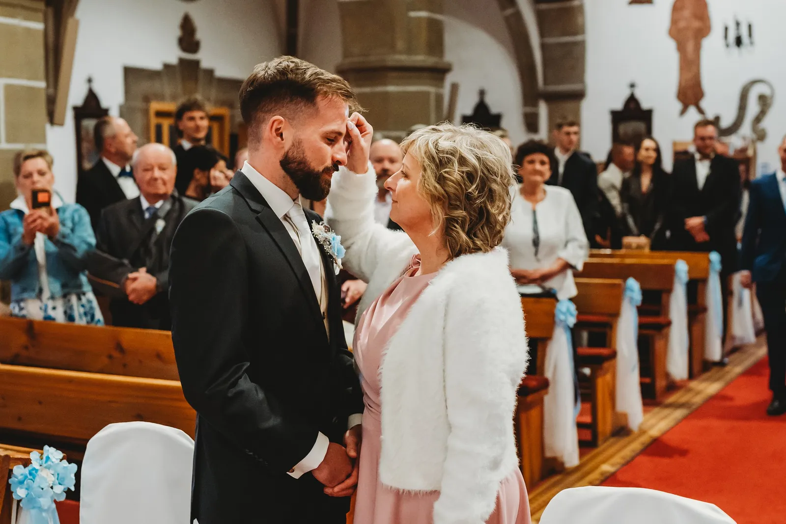 Mama v bielom svetríku žehná ženícha krížikom na čele pred oltárom v kostole – svadba Natália a Tomáš, Hotel Demänová