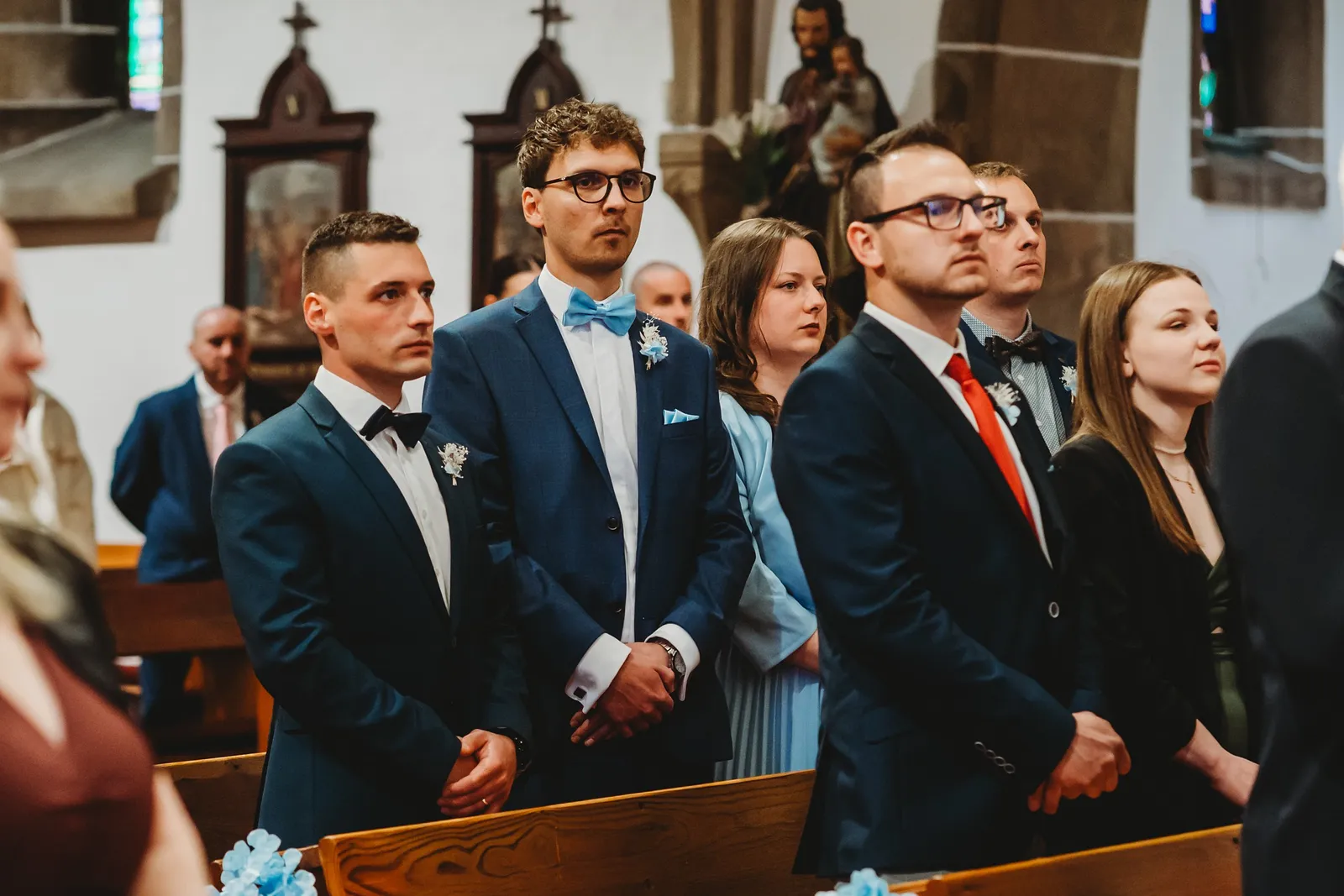 Svedkovia a hostia v tmavomodrých oblekoch stoja v kostolnej lavici počas obradu – svadba Natália a Tomáš, Hotel Demänová