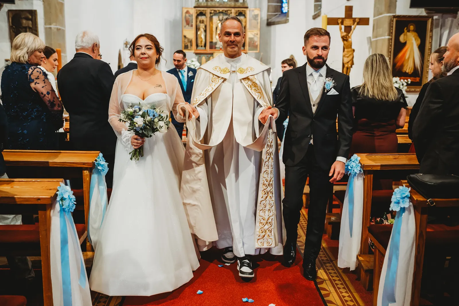 Kňaz vedie novomanželov uličkou z kostola, nevesta drží kyticu bielych ruží – svadba Natália a Tomáš, Hotel Demänová