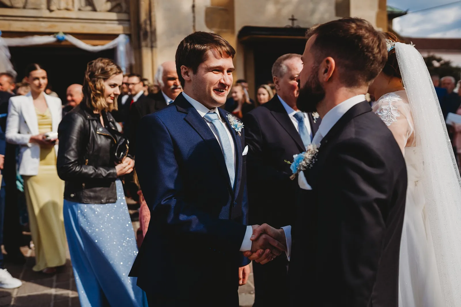 Hosť si podáva ruku so ženíchom pri gratuláciách pred kostolom na slnku – svadba Natália a Tomáš, Hotel Demänová