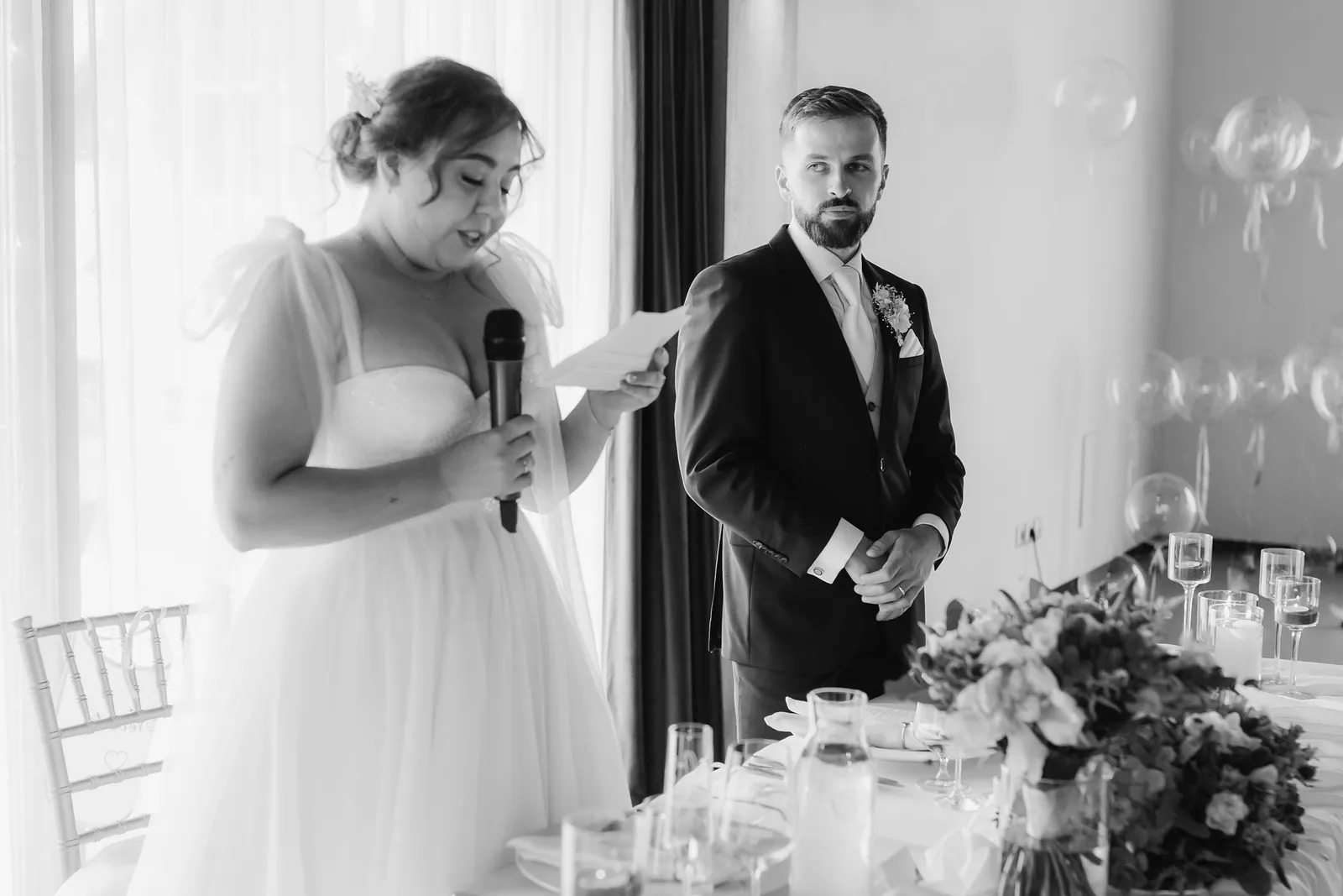 Nevesta s mikrofónom číta príhovor pri hodovom stole, ženích ju sleduje, čiernobiela snímka – svadba Natália a Tomáš, Hotel Demänová