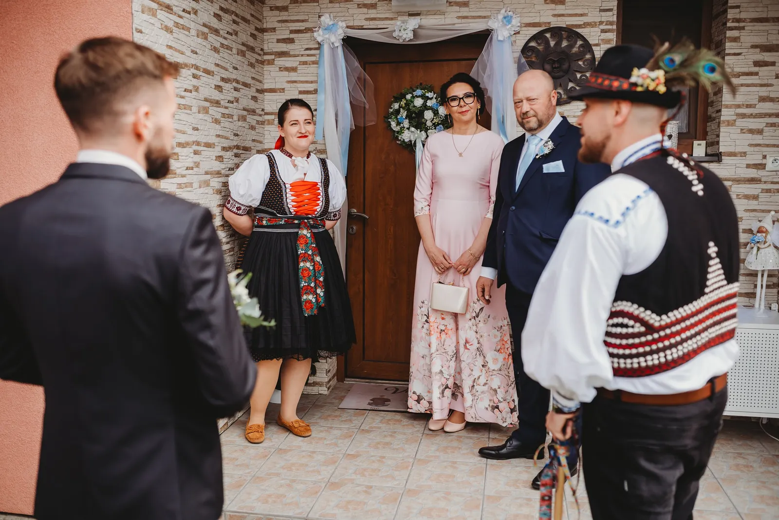 Ženích prichádza pred dom nevesty, kde ho čakajú rodičia, žena v kroji a starejší s klobúkom – svadba Natália a Tomáš, Hotel Demänová